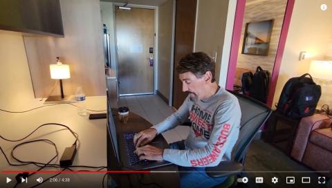 Screenshot from YouTube video of Bruce Wawrzyniak at desk in hotel on laptop