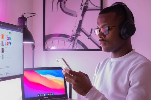 Male wearing headphones looking at phone in front of two computer screens
