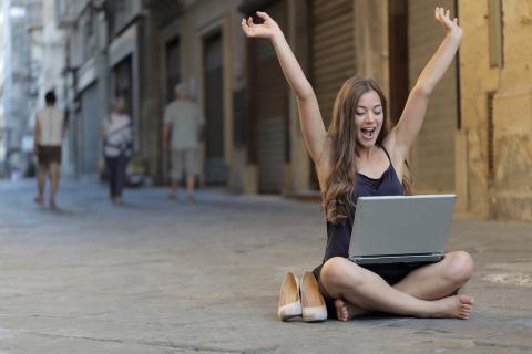 Girl sitting in street with laptop on knees while raising arms excitedly