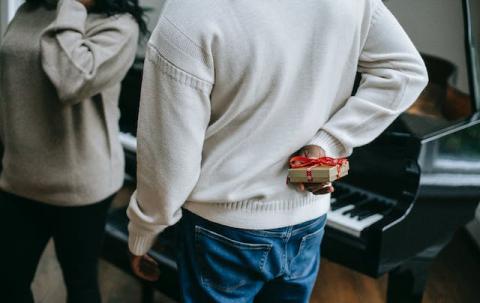 Male hiding small wrapped gift behind his back while standing next to a piano