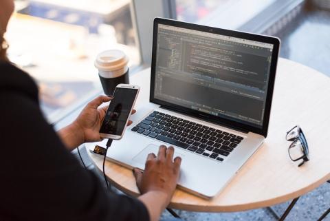 A female hand holds a smartphone with the other hand on a laptop keyboard with the screen displaying data