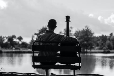 View from behind of guy sitting next to his guitar on a bench overlooking water