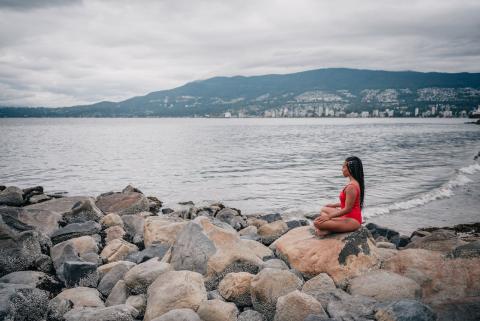A girl sits cross-legged on rocks alongside a lake