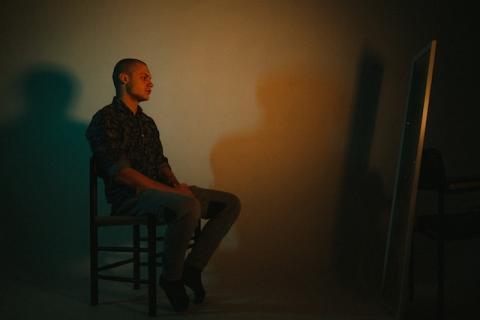 Man sitting on stool staring into floor length mirror in dark room
