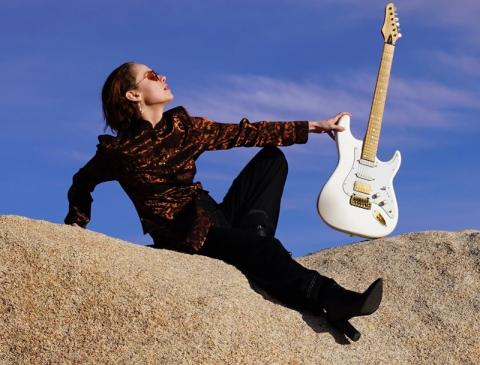 Raine Stern sits outside dressed in black wearing sunglasses holding an electric guitar