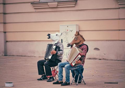 Two males seated on chairs on the sidewalk each playing an accordion while wearing a horse head mask