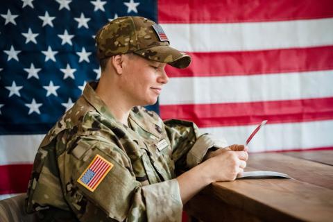 A solider in uniform sits in front of an American flag while looking reflectively at a photo