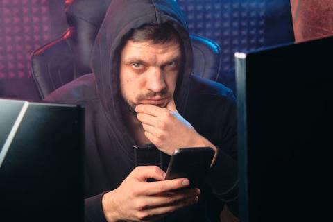 Male with hood up looking at cellphone while next to computer screen