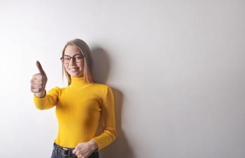 Girl indoors against plain wall wearing yellow shirt and glasses giving thumbs up