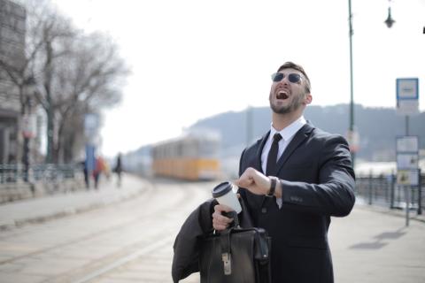 A man outdoors in a suit looks up frustratedly with his arm raised to see his watch