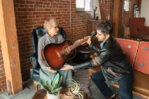 A middle aged man sits next to and helps a woman who is holding a guitar