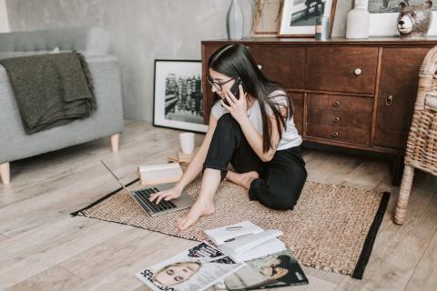 Girl talking on phone sits on floor looking at laptop with magazines nearby